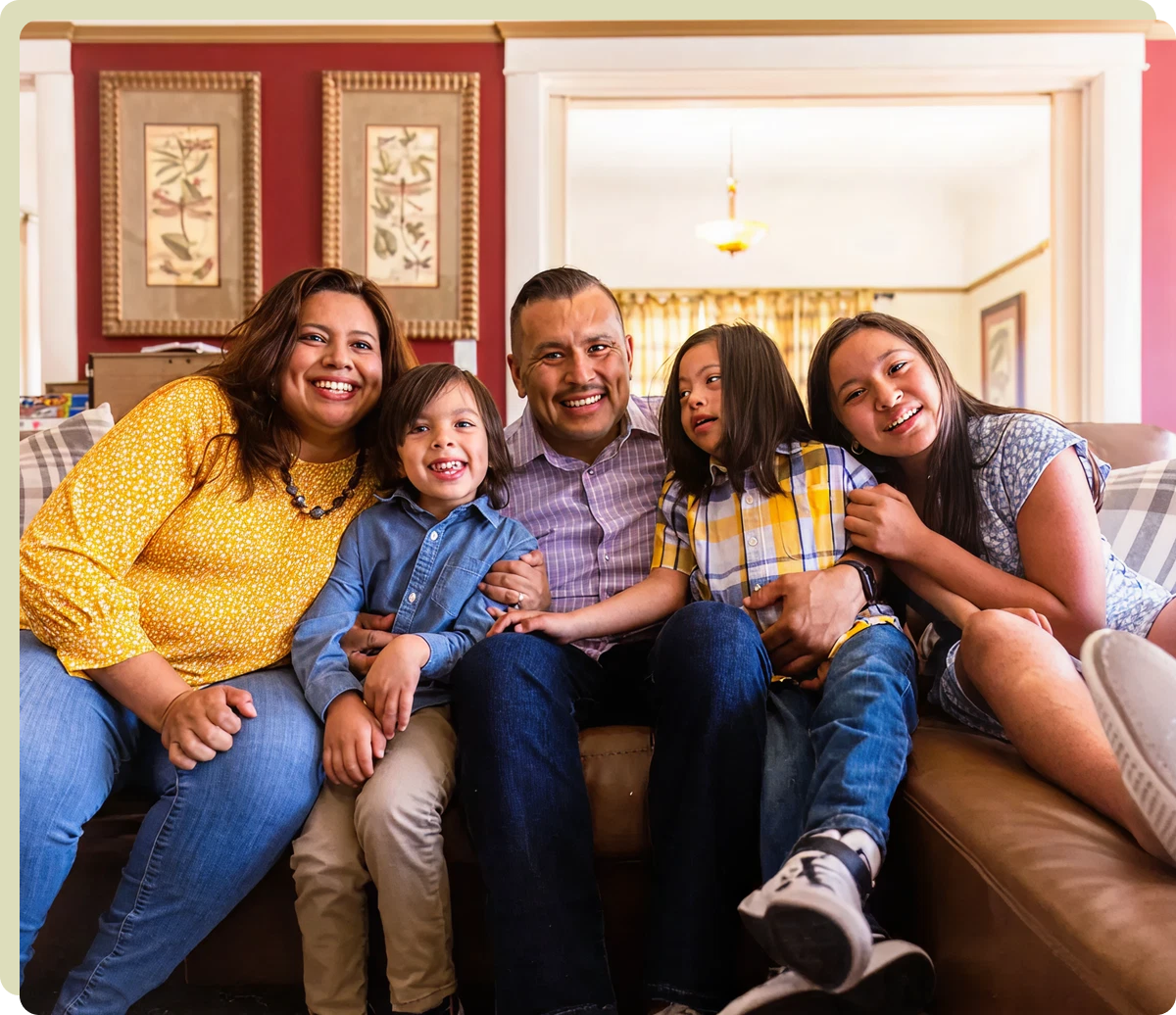 A family of five sits together on a couch, smiling at the camera in a warmly decorated living room.