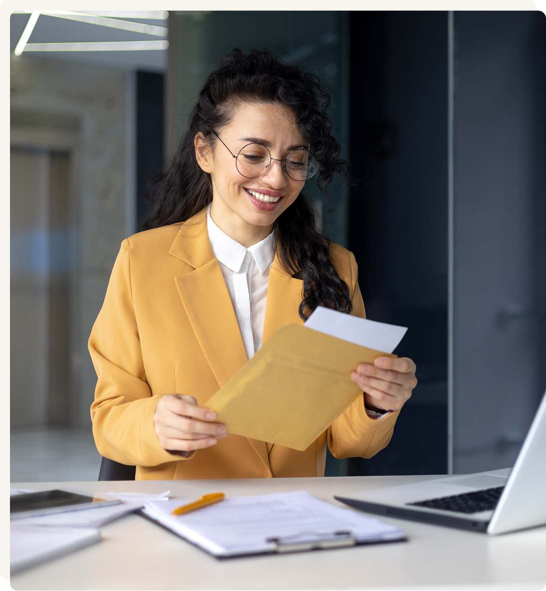 A woman wearing glasses and a yellow blazer smiles while opening a brown envelope at her desk, with documents, a pen, and a laptop in front of her.