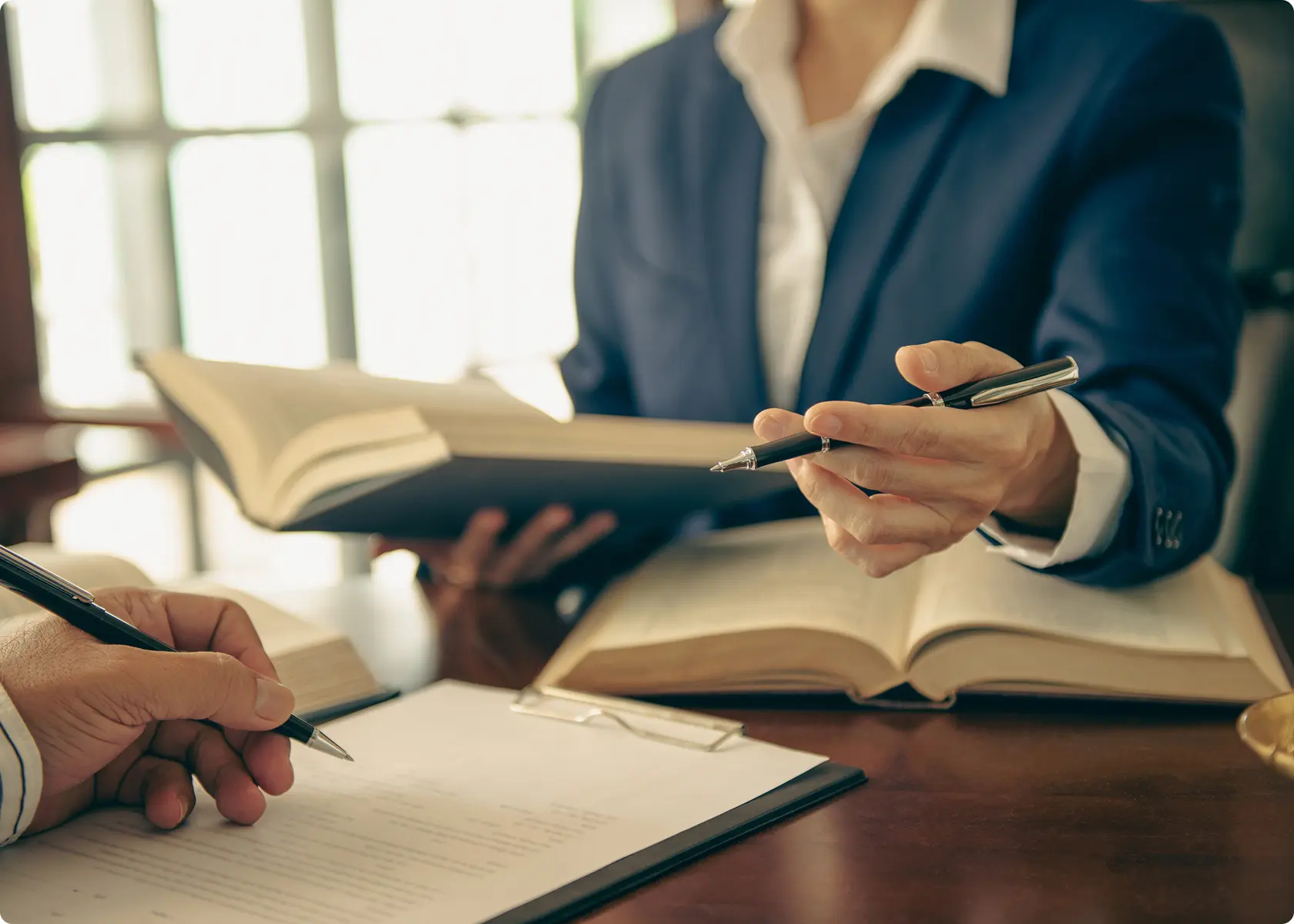Two people in business attire sit at a desk with open books; one points with a pen while the other writes on a clipboard.