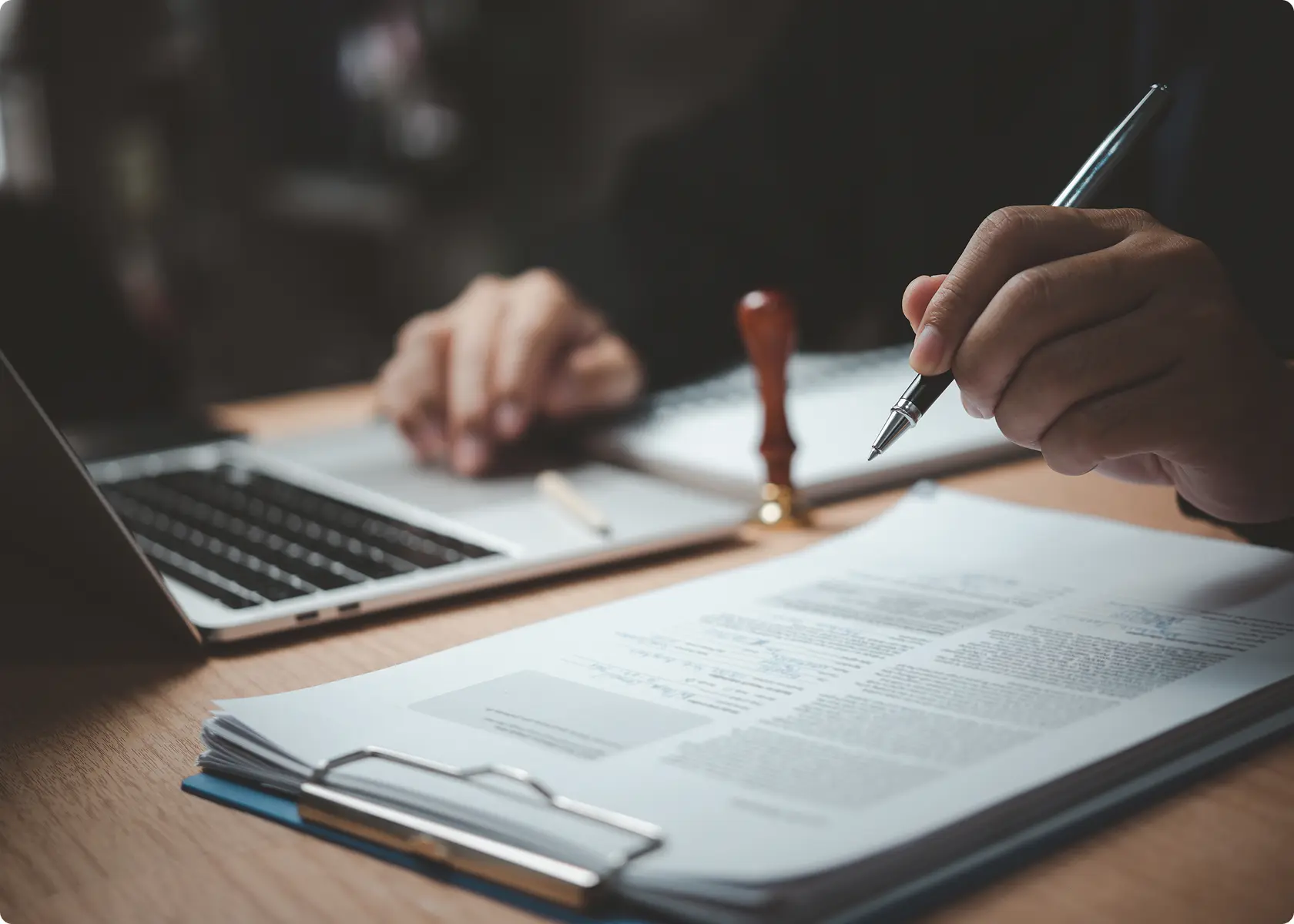 A person reviews and signs documents on a clipboard at a desk, with a laptop, notepad, pen, and a wooden stamp visible.