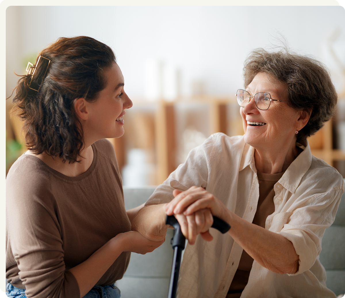 A younger woman smiles at an older woman seated with a cane; they hold hands and appear to be having a pleasant conversation indoors.