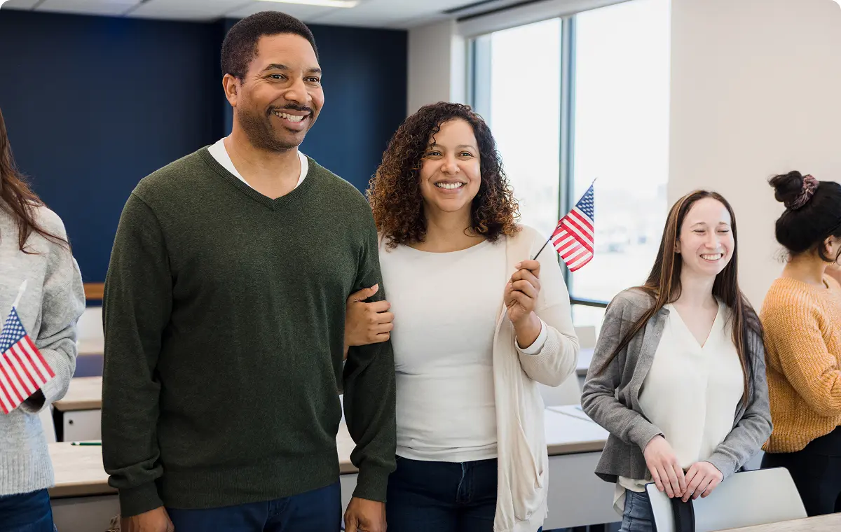A smiling couple stands arm-in-arm at an indoor event, with the woman holding a small American flag; others holding flags are visible in the background.