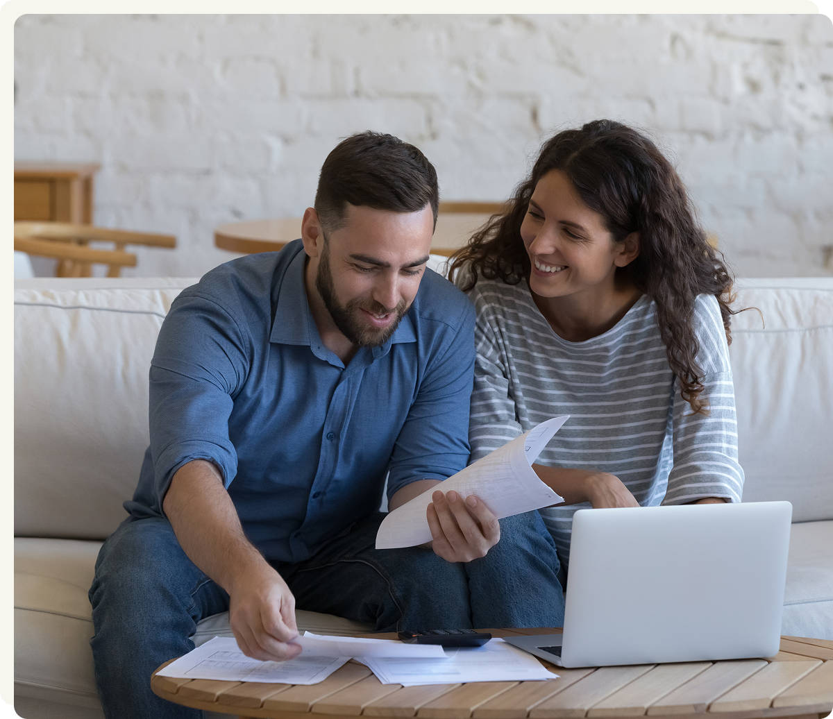 A man and woman sit on a couch, looking at papers and a laptop on a coffee table, both smiling and appearing to discuss something together.