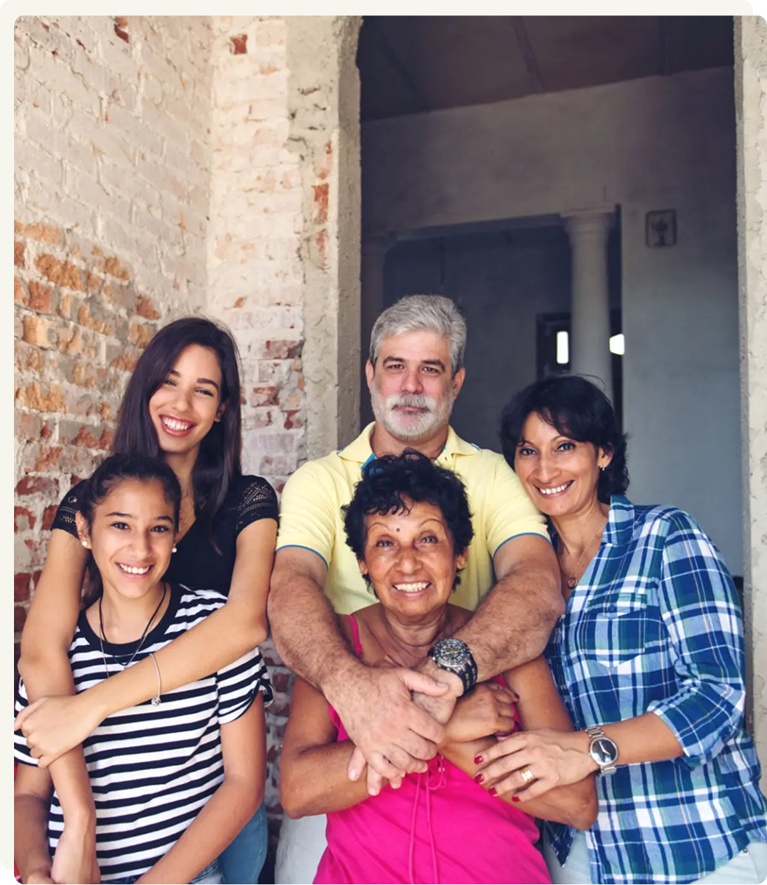 Five people, spanning three generations, stand closely together smiling in front of a rustic brick wall and doorway, suggesting a family portrait.